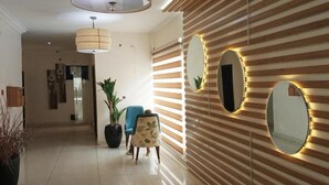 A modern lobby with a tiled floor, two large ceiling lights, a potted plant, a blue chair, and a wall with striped panels and three mirrors. - Nevada Hotels and Suites Lekki (Lekki)