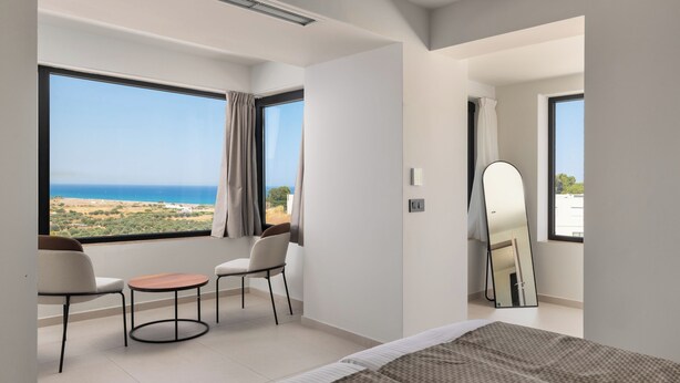 A bedroom with a bed, two chairs, a small table, a mirror, and large windows with a view of the sea. - Trapezia Luxury Living - Turquoise (Afantou)