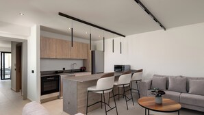A modern kitchen with a central island, bar stools, a grey sofa, and a round coffee table. - Trapezia Luxury Living - Turquoise (Afantou)