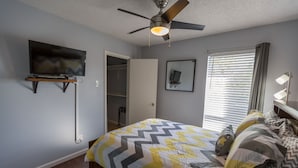 Un dormitorio con cama, ventilador de techo, televisor montado en la pared, ventana con persianas y mesita de noche con lámpara.