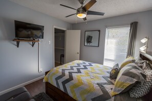 A bedroom with a bed, a ceiling fan, a TV mounted on the wall, a window with blinds, and a bedside table with a lamp.
