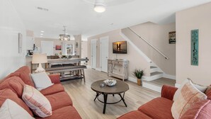Un vaste salon comprenant un grand canapé, une table basse ronde et un coin repas avec une table et des chaises en bois. Un escalier avec une rampe blanche mène à un autre niveau.