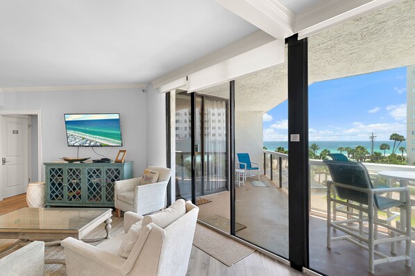A living room with a TV, a sofa, a coffee table, and a balcony with a view of the ocean.