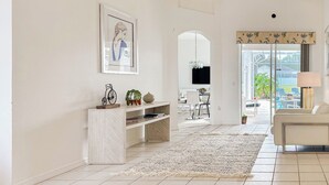 Un salón amplio con un sofá blanco, una mesa de centro y un televisor montado en la pared. Hay una estantería con objetos decorativos y un ventanal con vistas al exterior.