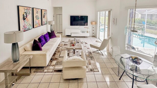 Una sala de estar moderna con un sofá, una mesa de centro y un televisor. Hay una mesa de vidrio con sillas y vistas a una piscina.