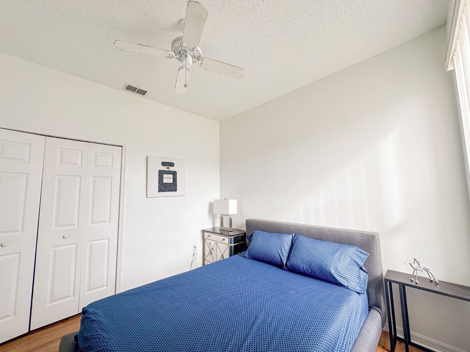 A bedroom with a bed, a ceiling fan, a bedside table with a lamp, and a framed picture on the wall.