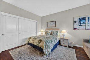 Un dormitorio con una cama, dos mesitas de noche, una mesita auxiliar, una puerta y una pared con un cuadro.