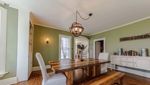 A dining area with a wooden table, white chairs, a chandelier, and a farmhouse gathering sign. - The 1840's Goat House (Elburn)