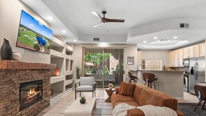 A modern living room with a fireplace, a large flat-screen TV mounted on the wall, a ceiling fan, and a kitchen area with stainless steel appliances. - Single Level on TPC Champions Course | 2 Bedroom (Scottsdale)