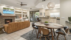A modern living room with a fireplace, a large TV, and a dining area with a round table and chairs. - Single Level on TPC Champions Course | 2 Bedroom (Scottsdale)