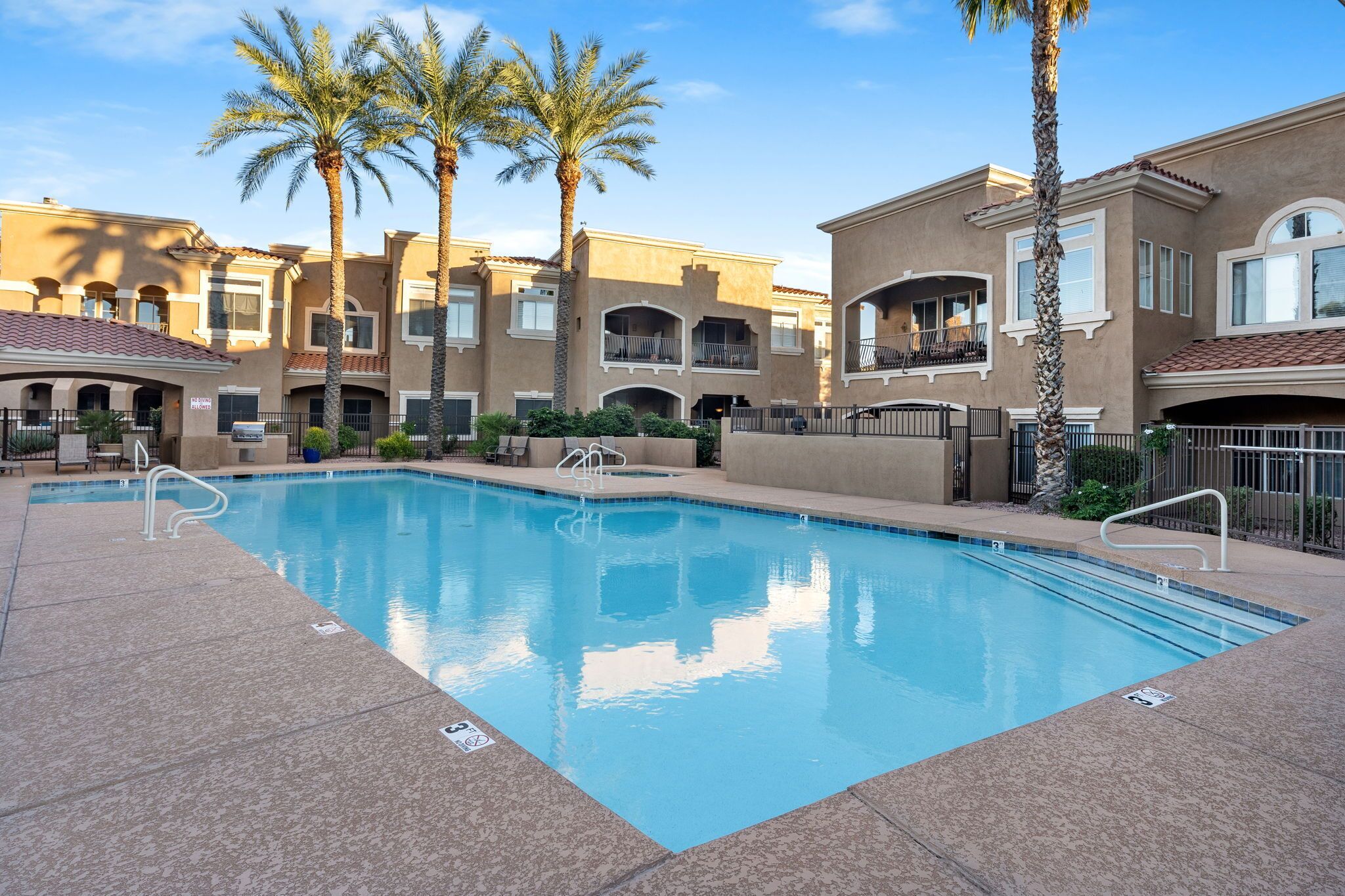 A swimming pool with a tiled deck, surrounded by multi-story buildings with balconies and palm trees.