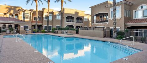 A swimming pool with a tiled deck, surrounded by multi-story buildings with balconies and palm trees.