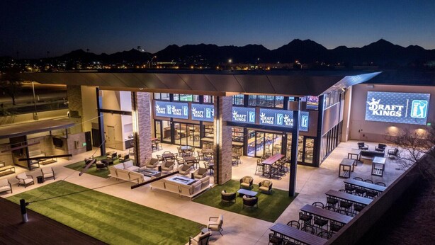 A modern outdoor dining area with multiple seating arrangements, a large screen displaying 'Draft Kings', and a view of mountains in the background. - Single Level on TPC Champions Course | 2 Bedroom (Scottsdale)