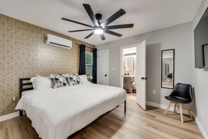 A bedroom with a bed, a ceiling fan, an air conditioning unit, a wooden floor, and a door leading to a bathroom.