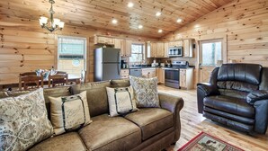 Un acogedor interior de cabaña con techo de madera, una zona de comedor con mesa y sillas, una cocina con electrodomésticos y un sillón de cuero reclinable.
