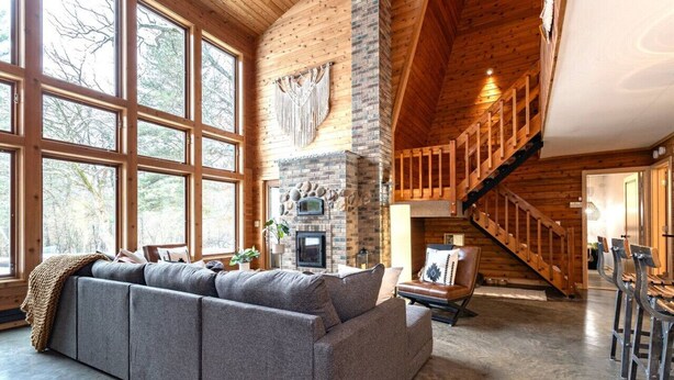 A spacious living area with a large window, a stone fireplace, a wooden staircase, and a grey sofa. - Rapids Rustic Retreat (Wisconsin Rapids)