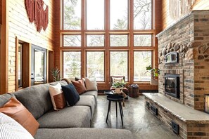 A cozy living room with a fireplace, large windows, and a wooden interior. - Rapids Rustic Retreat (Wisconsin Rapids)