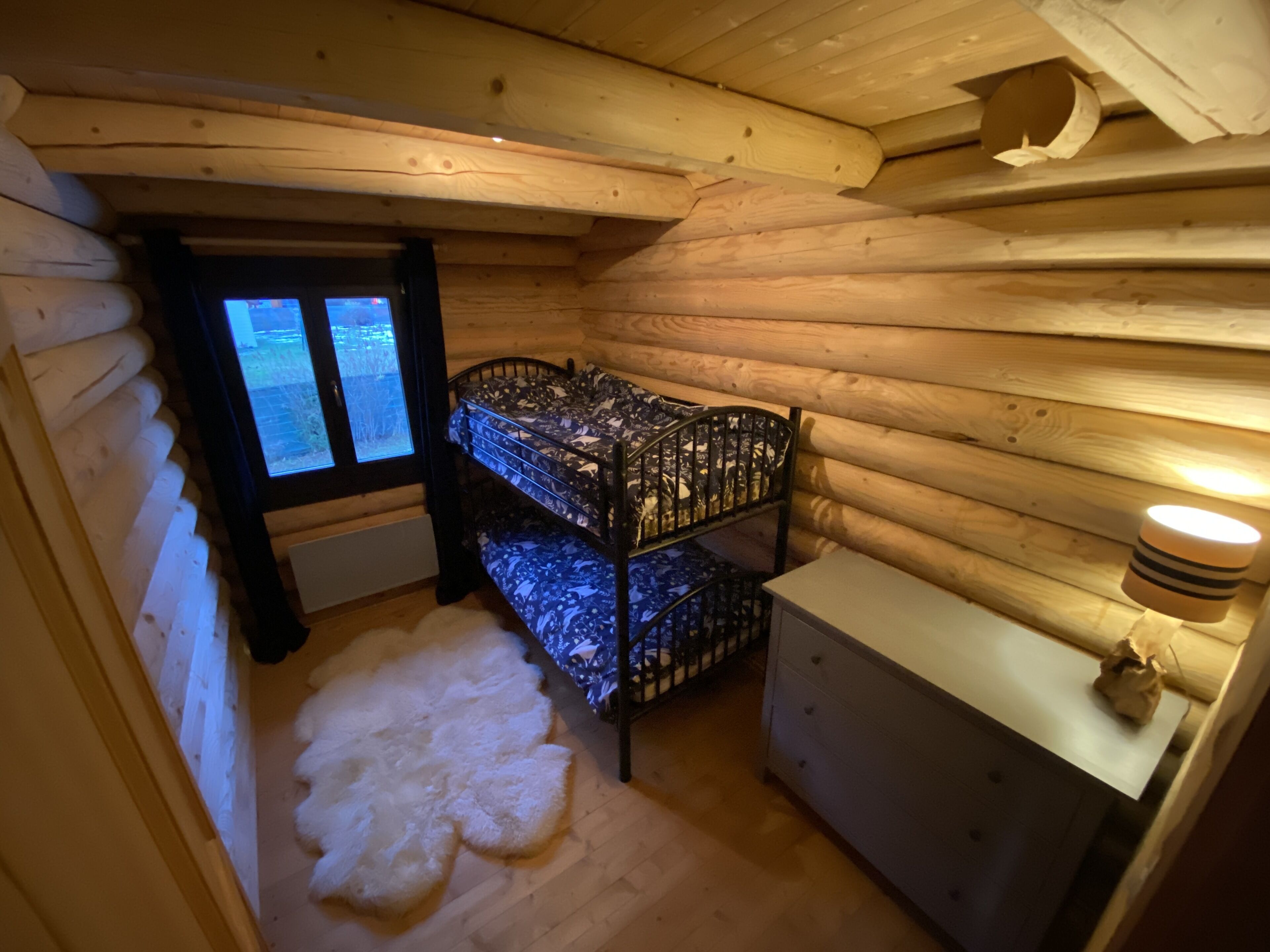 A cozy cabin room with a bunk bed, a white fur rug, a wooden dresser, and a window with a view of the outdoors.