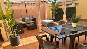Un patio con una mesa de vidrio, sillas de mimbre, un sofá con cojines y plantas en macetas.