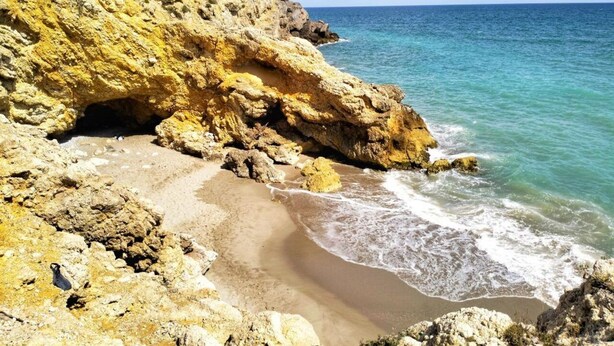 Paisaje costero con acantilados rocosos, una playa de arena y mar azul cristalino.