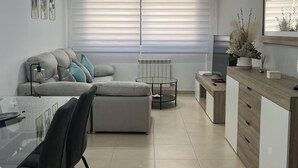 Una sala moderna con un sofá gris, una mesa de comedor de vidrio y una mesa de centro. Hay un radiador debajo de las persianas.