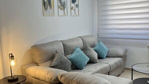 Una sala de estar con un sofá gris, cojines color turquesa, una mesita auxiliar con una lámpara, y arte botánico enmarcados en la pared.