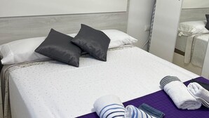 Una cama bien hecha con una colcha blanca y morada, dos almohadas y una toalla doblada sobre una manta morada.