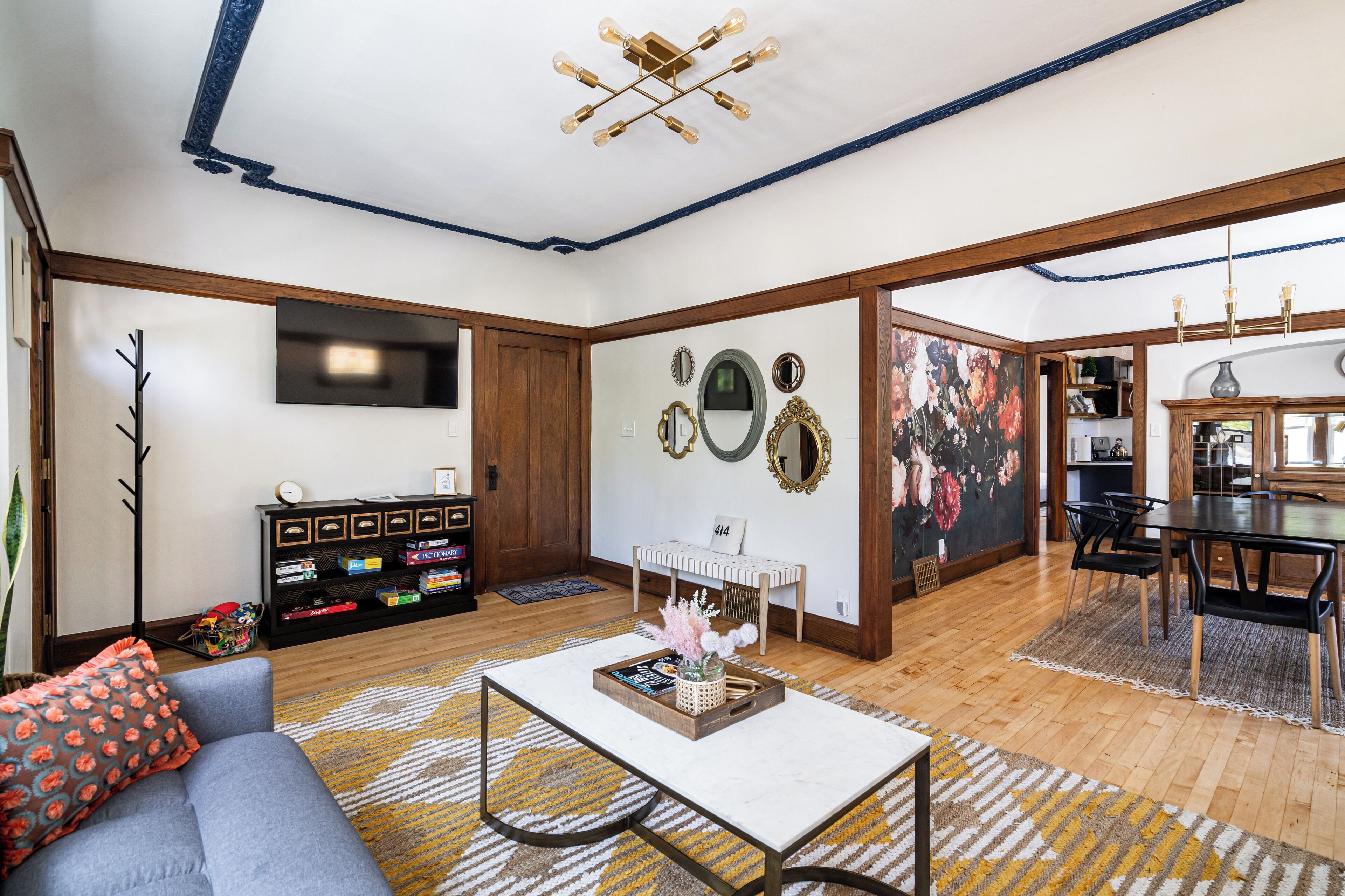 A spacious living room with a sofa, coffee table, and a dining area with a table and chairs. There is a TV mounted on the wall, a bookshelf, and a decorative mirror.