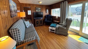 Un salon chaleureux avec un plancher en bois, un canapé, un fauteuil, un téléviseur et une fenêtre donnant sur une maison.