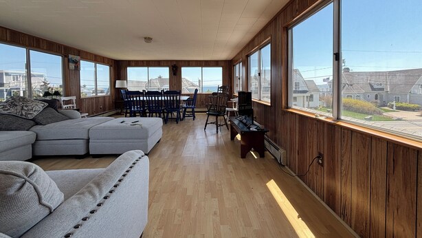 Un vaste salon comprenant un grand canapé d’angle, une table à manger avec des chaises et un piano. Les murs de la pièce sont recouverts de lambris en bois et de grandes fenêtres offrent une vue sur le quartier.