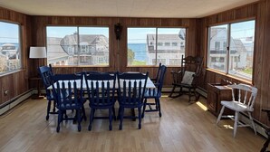 Un coin repas avec une table et des chaises en bois, de grandes fenêtres donnant sur des maisons et la mer, et un luminaire au plafond.