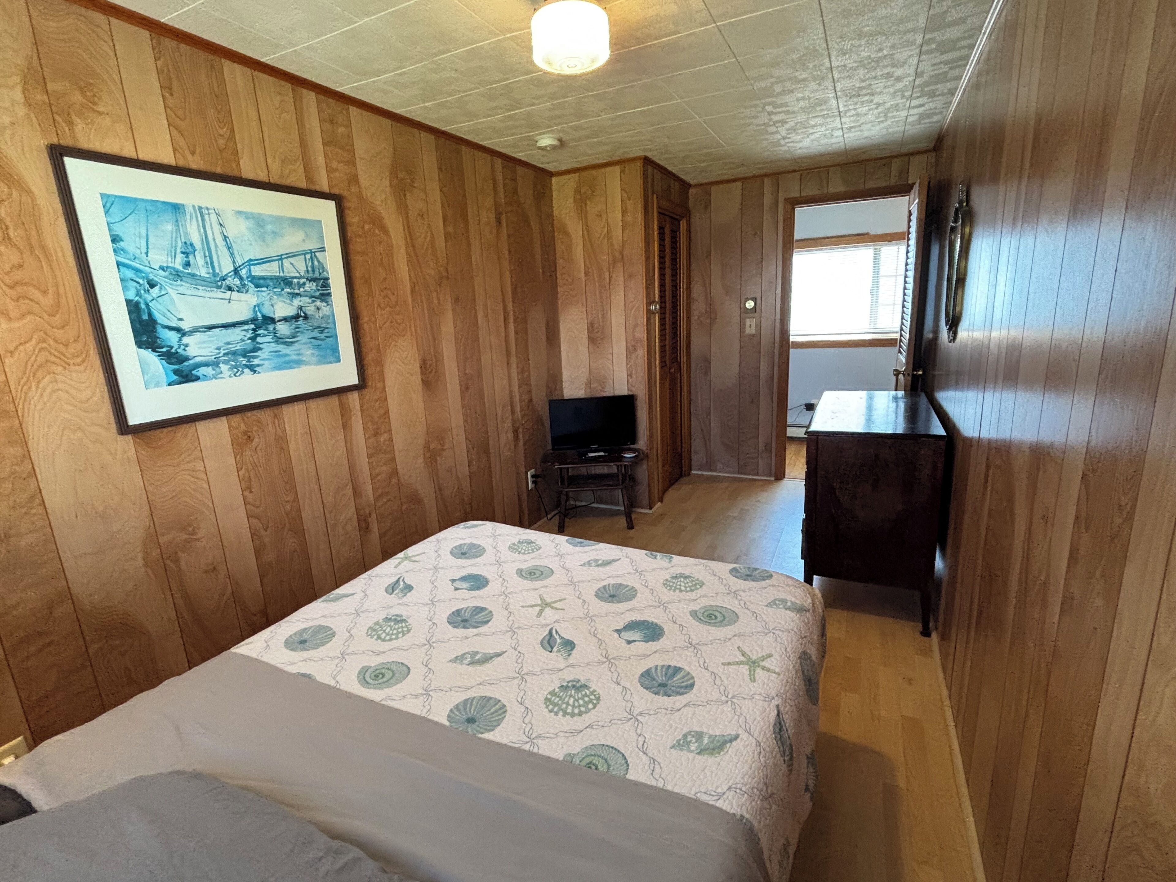Une chambre avec des lambris en bois, un lit recouvert d’une couvre-lit à motifs, un téléviseur et un tableau encadré accroché au mur.
