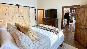 A bedroom with a bed, pillows, a wooden headboard, a television, and a kitchen area visible through an open door. - NEW! LAST MIN SPECIAL! Ski Chalet, Spacious, Clean, Close To Everything! (Big Sky)
