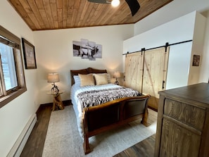 Een slaapkamer met een houten plafond, een bed met hoofdbord, een nachtkastje met een lamp, een kledingkast en een ingelijst schilderij aan de muur.