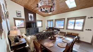 A cozy living room with a fireplace, wooden dining table, and large windows offering a view of the mountains. - NEW! LAST MIN SPECIAL! Ski Chalet, Spacious, Clean, Close To Everything! (Big Sky)