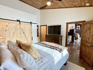 A bedroom with a bed, pillows, a wooden headboard, a television, and a kitchen area in the background. - NEW! LAST MIN SPECIAL! Ski Chalet, Spacious, Clean, Close To Everything! (Big Sky)