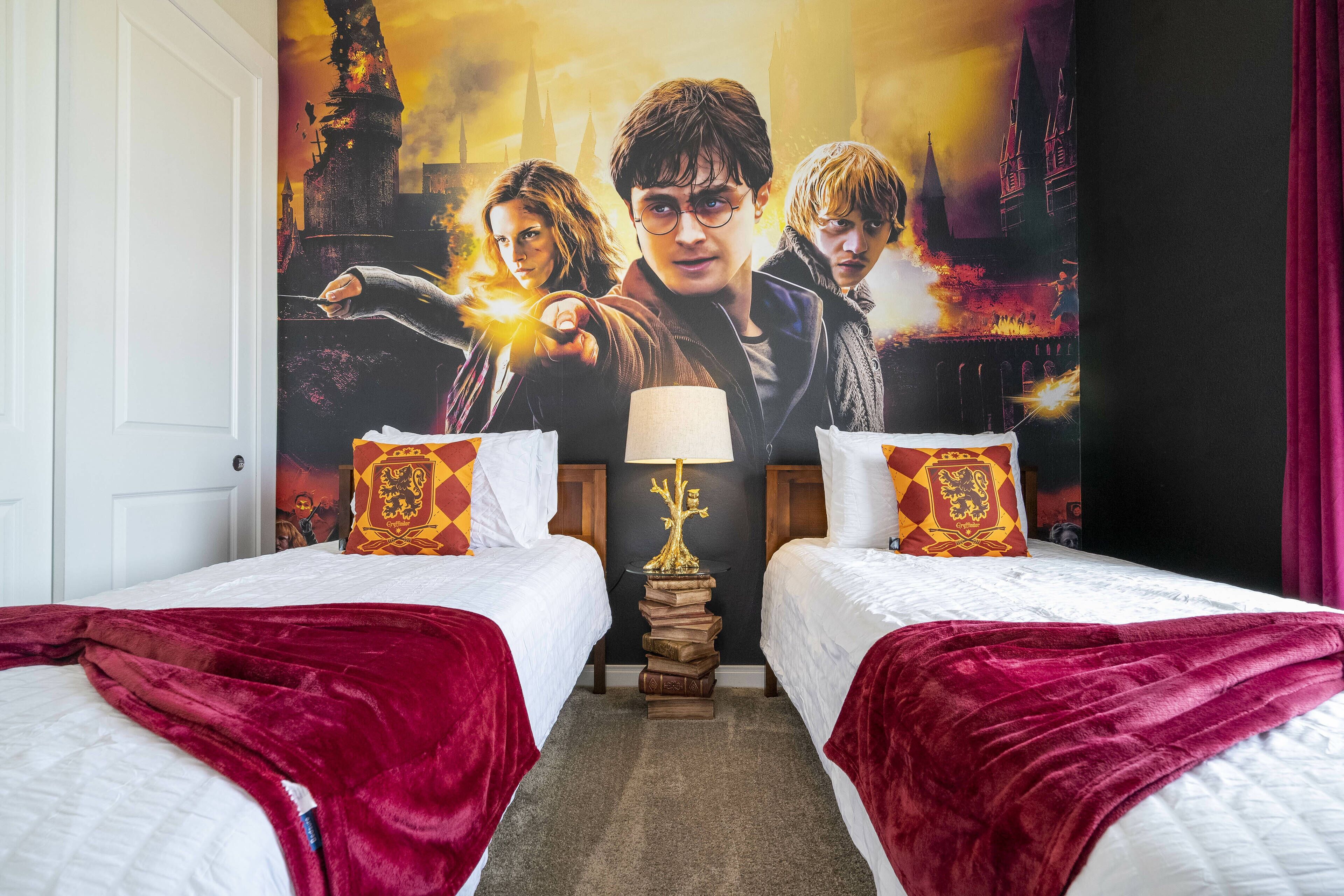 A hotel room with two beds, each with a red blanket and matching pillows. There is a wall-mounted lamp with a tree design on a stack of books.