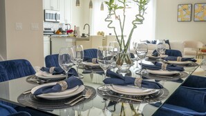 A dining area with a glass table set for six, blue upholstered chairs, and a vase with green plants. - 2024 Unique Villa 5 min to Disney (Kissimmee)