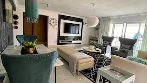 A modern living room with a sofa, a TV mounted on the wall, and a dining area with a table and chairs. - Granville Penthouse Apartments (Lekki)