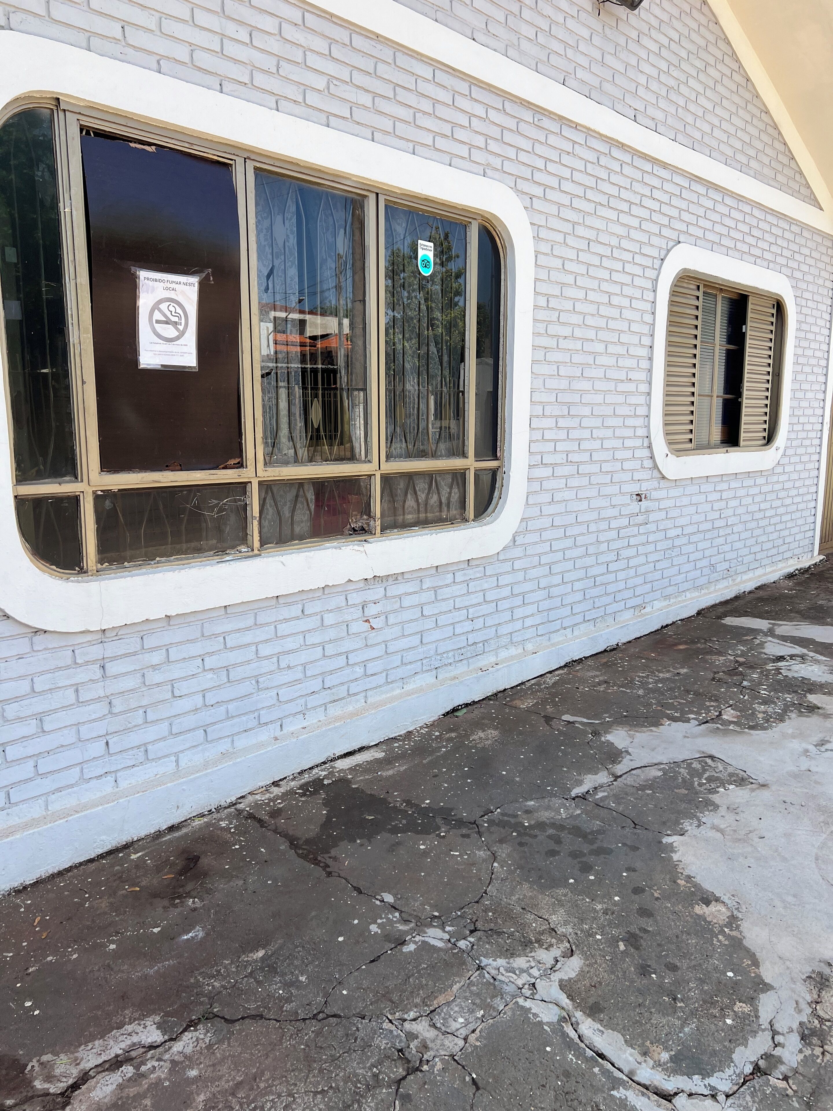 A building with a white brick exterior, large windows with metal bars, and a cracked pavement.