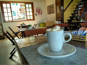 A white coffee cup on a saucer with a woven placemat, a wooden table, and a view of a dining area with chairs and a window.