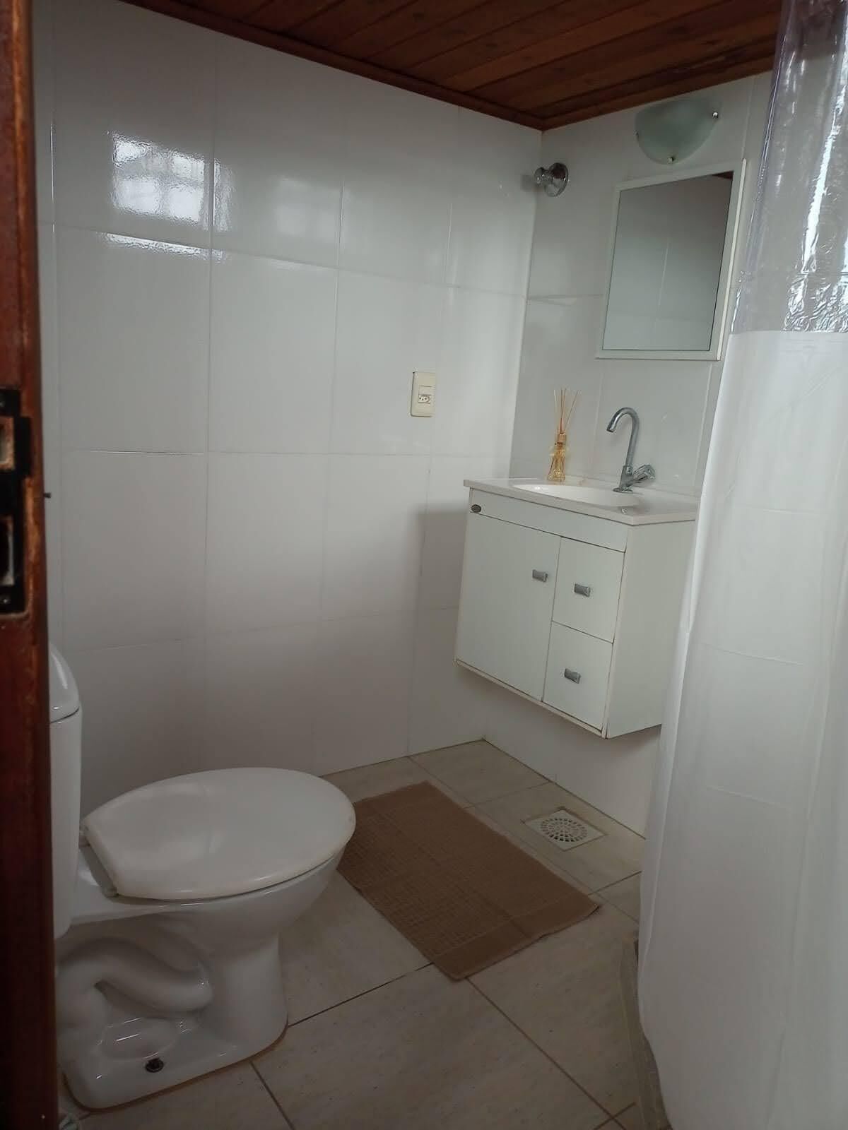 Un baño pequeño y limpio con azulejos blancos, un inodoro, un lavabo con espejo y una cortina de ducha.