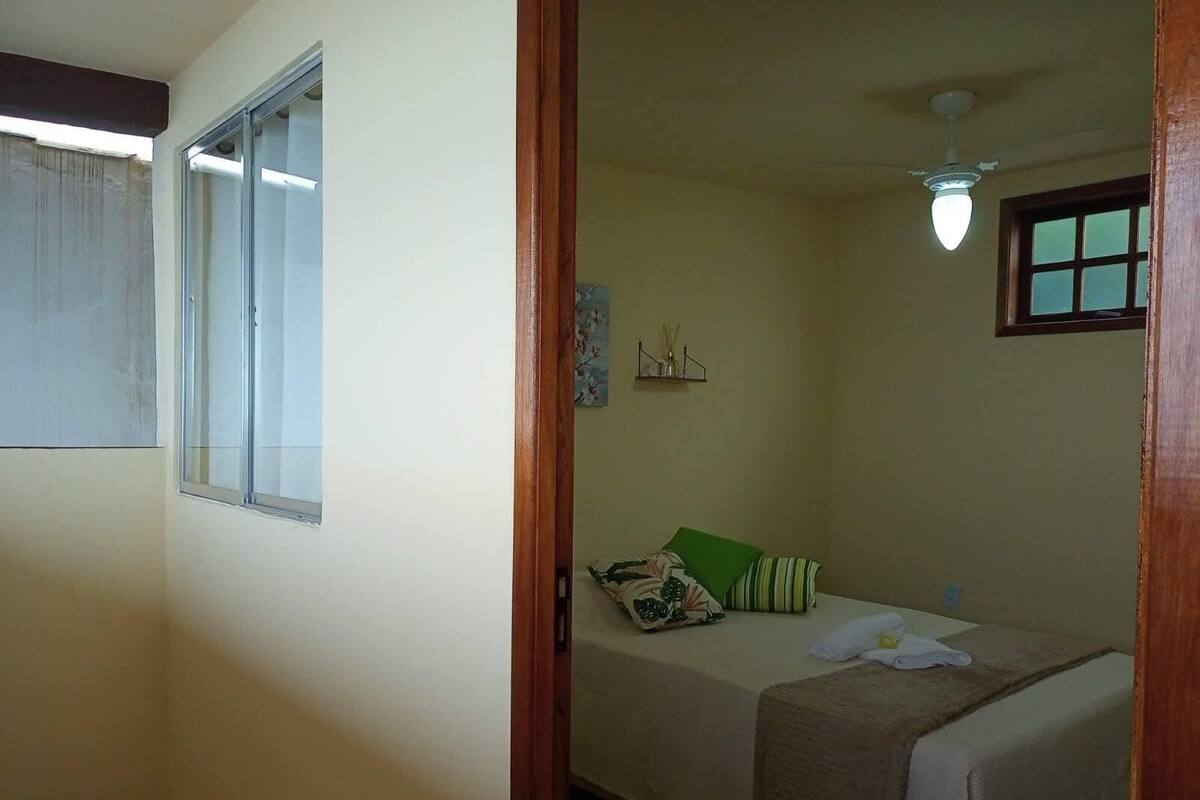 A small, single-bed room with a window, a ceiling fan, and a wall-mounted shelf with decorative items.