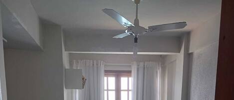 Una habitación pequeña y limpia con dos camas, un ventilador de techo, una ventana con cortinas y una pequeña mesa con una silla.