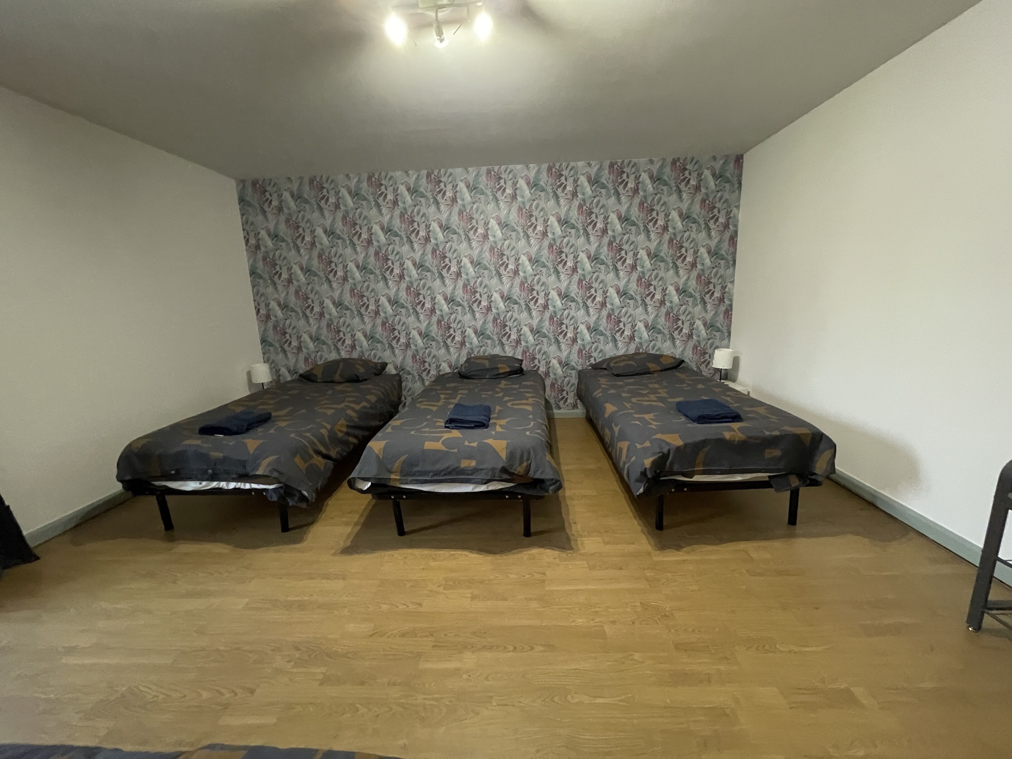 A room with three single beds, a patterned wallpaper, and wooden flooring.