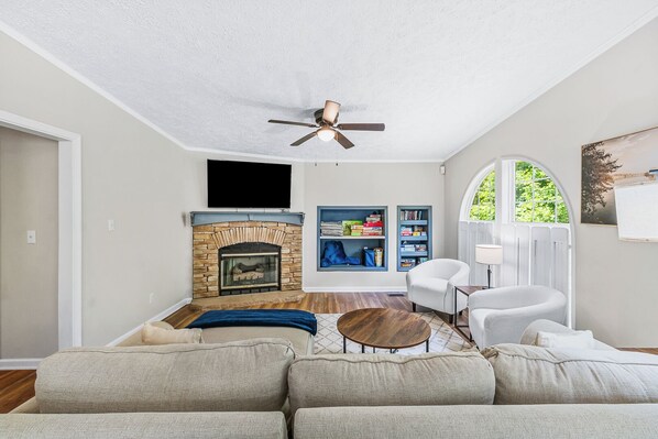 A living room with a fireplace, a ceiling fan, a bookshelf, and a large window.