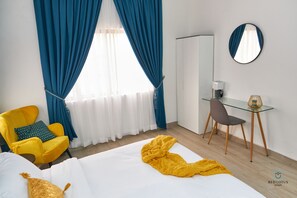 A hotel room with a bed, a yellow armchair, a desk with a chair, a round mirror, and a window with blue curtains. - Cheerful Modern Studio Apartment in Ghana with Fast WIFI & Stunning City Views.  (Accra)
