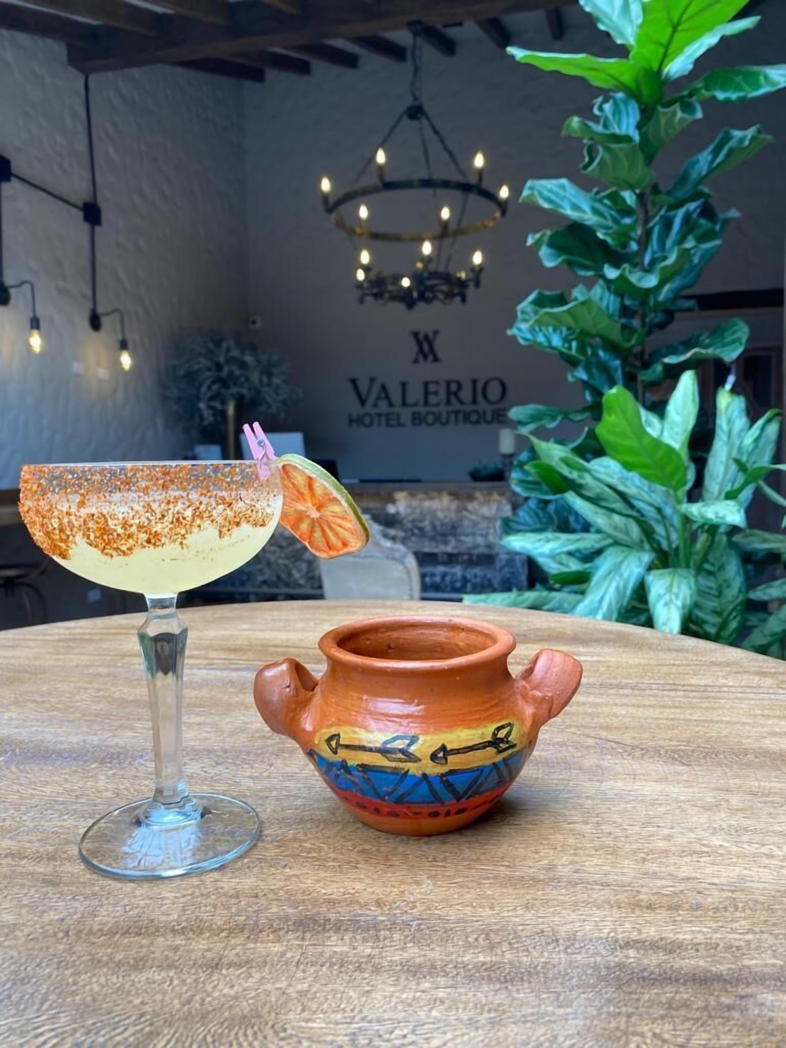 A cocktail glass with a salted rim and a slice of orange on the rim, a small clay pot with colorful designs, and a sign for 'Valerio Hotel Boutique' in the background.