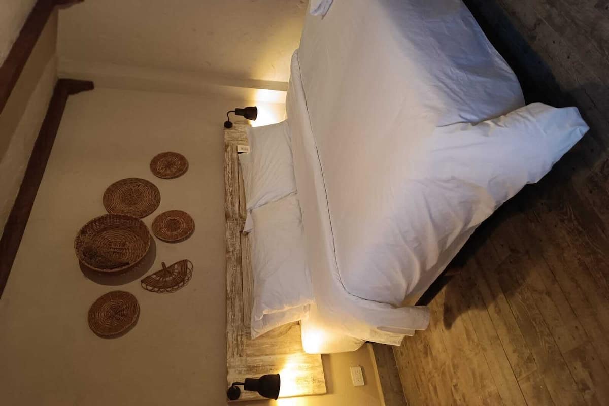 A neatly made bed with white linens, two wall-mounted lamps, and woven wall art.