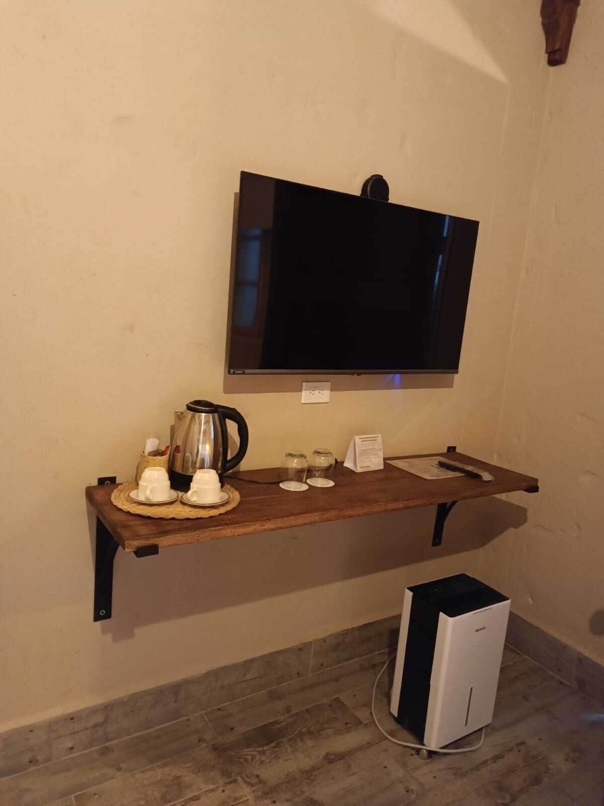 Una habitación con una televisión de pantalla plana montada, un estante de madera con una cafetera, tazas y un menú, y una consola de videojuegos blanca en el suelo.